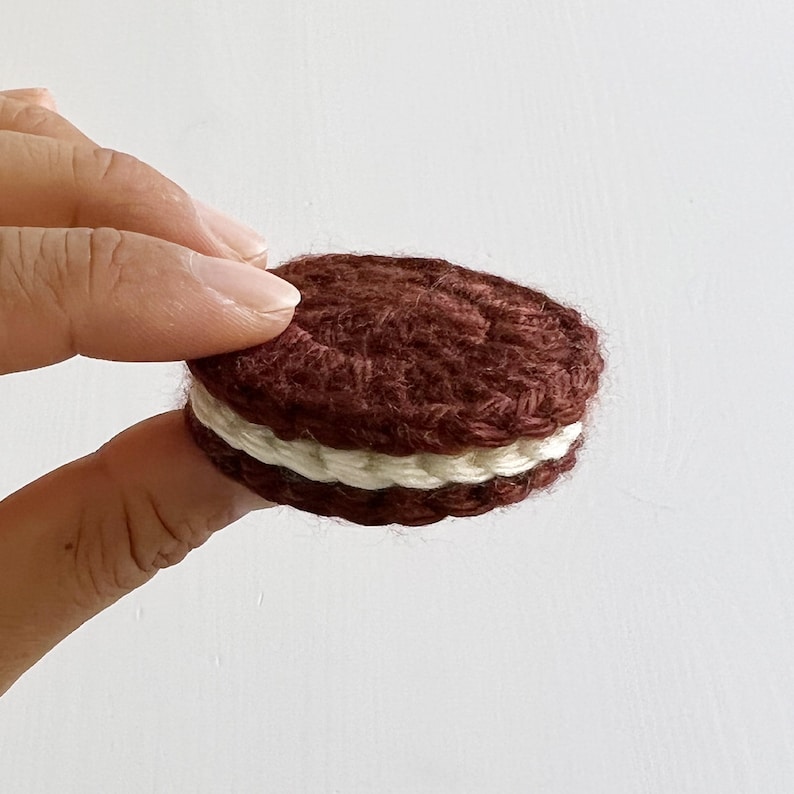 May include: A crocheted chocolate sandwich cookie with white frosting. The cookie is brown and has a textured surface.