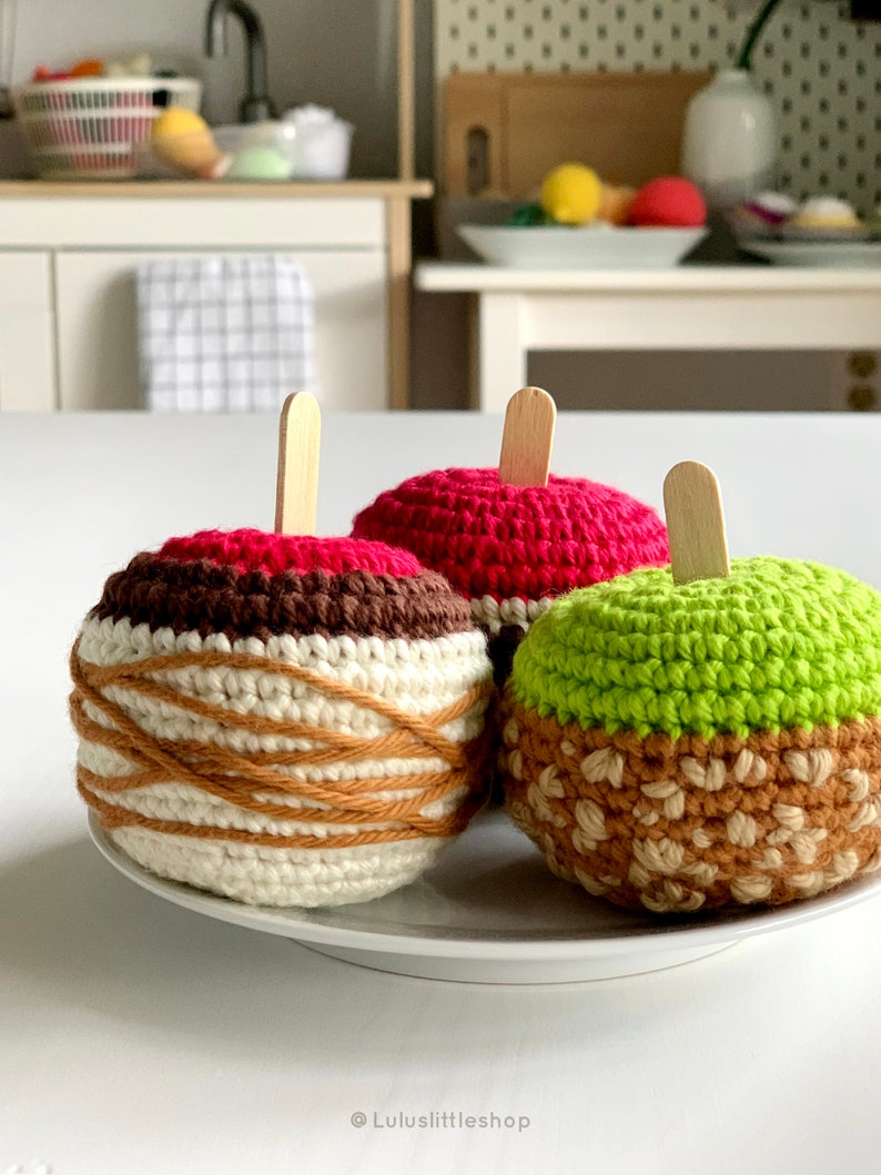 Puede incluir: Tres juguetes de ganchillo con forma de manzana con palitos de madera para los tallos. Las manzanas son rojas, verdes y blancas con detalles marrones. Las manzanas est&aacute;n en un plato blanco.