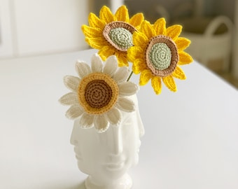 Puede incluir: Tres girasoles de crochet en un jarrón blanco con un diseño de cara. Los girasoles son amarillos con centros marrones y detalles verdes. El jarrón es una pieza única con múltiples caras.