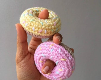Op de afbeelding: Twee gehaakte donuts, een gele met roze en blauwe sprinkles en een roze met gele en blauwe sprinkles.
