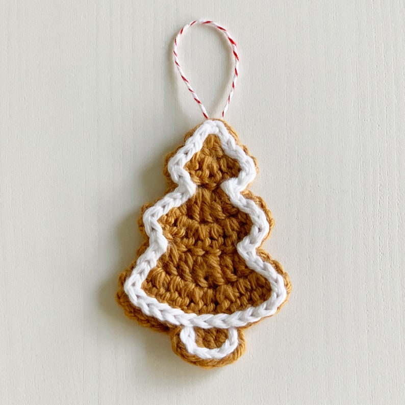K&ouml;nnte beinhalten: Ein geh&auml;kelter Lebkuchenmann-Anh&auml;nger in Form eines Weihnachtsbaums mit wei&szlig;en Zuckergussdetails. Der Anh&auml;nger ist braun und wei&szlig; und hat eine rot-wei&szlig;e Schnur zum Aufh&auml;ngen.