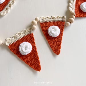 May include: A crocheted pumpkin pie garland with orange and white fabric. The garland is strung with wooden beads and features a white crocheted flower on each pumpkin pie.
