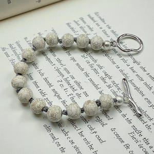 May include: A beaded bracelet featuring off-white, patterned beads strung on a gray cord. The bracelet has a silver-toned toggle clasp and is displayed on an open book with aged pages.