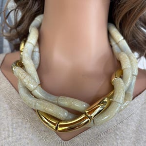 May include: A statement necklace with large, cream-coloured, textured links and gold-tone accents. The necklace is made up of multiple strands and is worn around the neck. The gold-tone elements provide a contrast to the cream-coloured links.
