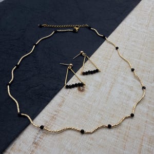 May include: A gold-toned necklace and matching earrings set. The necklace has a wavy design with black beads. The earrings are triangular with black beads. The jewelry set is displayed on a black and white surface.