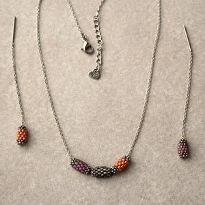 May include: A silver necklace and matching earrings set. The necklace features a beaded design with purple, gray, and orange beads. The earrings have a similar beaded design. The jewelry set is displayed on a neutral background.