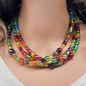 May include: A multi-strand necklace featuring colorful glass beads in shades of red, orange, yellow, green, blue, and purple. The beads are various shapes and sizes, creating a layered effect. The necklace is worn around the neck.