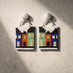 Op de afbeelding: Een paar kralen oorbellen met een ontwerp van een boodschappentas. De oorbellen hebben een rechthoekige vorm met een zilverkleurige metalen handgreep en stud. De tassen zijn wit, zwart, blauw, oranje en groen. Op een neutrale achtergrond.