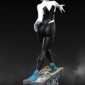 May include: A figurine of a female superhero in a black and white costume with blue footwear. The figure is posed on a stone structure with a spiderweb design. The statue has short blonde hair and is set against a dark background.