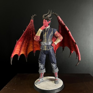Puede incluir: Figura detallada de un personaje masculino con piel roja, cuernos y grandes alas rojas. Viste un atuendo azul oscuro y dorado con detalles plateados. La figura se encuentra sobre una base redonda y embaldosada, con un fondo oscuro.