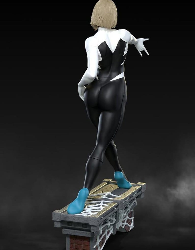 May include: A statue of a female superhero in a black and white suit with blue shoes. The figure is posed on a stone platform with a spiderweb design. The superhero has short blonde hair and is in a dynamic pose.