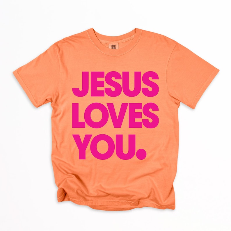 K&ouml;nnte beinhalten: Ein orangefarbenes T-Shirt mit der Aufschrift "JESUS LOVES YOU." in leuchtend pinkfarbenen Blockbuchstaben. Das Shirt hat kurze &Auml;rmel und einen Rundhalsausschnitt. Der Text ist mittig auf der Vorderseite des Shirts platziert.