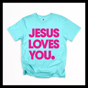 K&ouml;nnte beinhalten: Hellblaues T-Shirt mit dem Text "JESUS LOVES YOU." in leuchtend pinkfarbenen Gro&szlig;buchstaben. Das T-Shirt mit Rundhalsausschnitt ist aus weichem Stoff. Der Text ist mittig auf der Vorderseite des Kleidungsst&uuml;cks platziert.