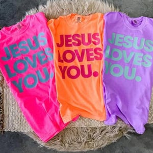 K&ouml;nnte beinhalten: Drei farbenfrohe T-Shirts: Pink, Orange und Lila. Jedes Shirt tr&auml;gt den Text "JESUS LOVES YOU" in kontrastierenden Farben. Die Shirts liegen auf einer strukturierten Oberfl&auml;che.