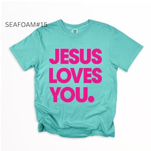K&ouml;nnte beinhalten: T&uuml;rkisfarbenes T-Shirt mit dem Text "JESUS LOVES YOU" in leuchtendem Pink. Das Shirt hat einen Rundhalsausschnitt und scheint aus einem weichen Material zu sein. Der Text "SEAFOAM#15" befindet sich in der oberen linken Ecke.