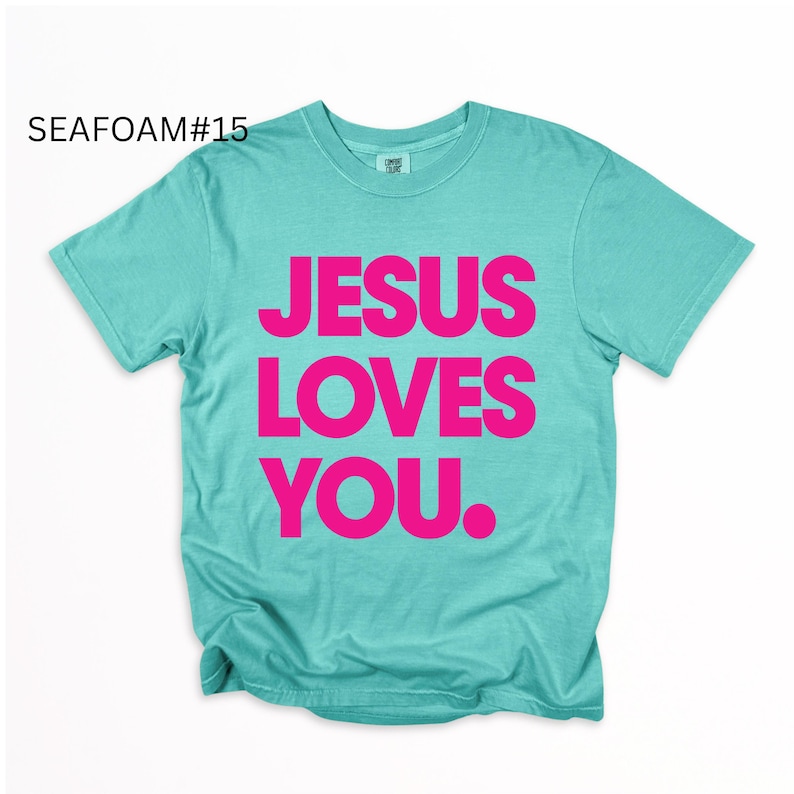 K&ouml;nnte beinhalten: T&uuml;rkisfarbenes T-Shirt mit dem Text "JESUS LOVES YOU" in leuchtendem Pink. Das Shirt hat einen Rundhalsausschnitt und scheint aus einem weichen Material zu sein. Der Text "SEAFOAM#15" befindet sich in der oberen linken Ecke.