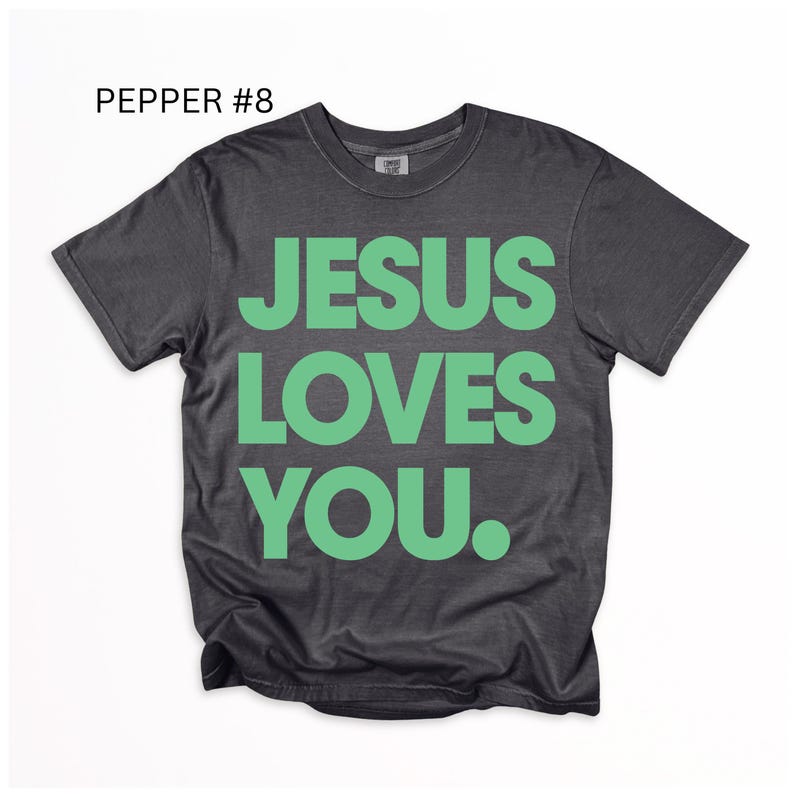 K&ouml;nnte beinhalten: Ein dunkelgraues T-Shirt mit dem Text "JESUS LOVES YOU" in gro&szlig;en, leuchtend gr&uuml;nen Buchstaben. Das Shirt hat auch den Text "PEPPER #8" in der oberen linken Ecke. Das Shirt ist ein Rundhals und hat kurze &Auml;rmel.