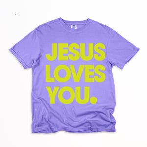 K&ouml;nnte beinhalten: Ein lilafarbenes T-Shirt mit dem Text "JESUS LOVES YOU" in leuchtendem Gelb. Das Shirt hat kurze &Auml;rmel und einen Rundhalsausschnitt und ist aus weichem Material gefertigt. Der Text ist mittig auf der Vorderseite platziert.