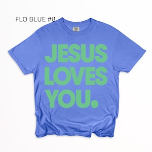 K&ouml;nnte beinhalten: Ein blaues T-Shirt mit dem Text "JESUS LOVES YOU" in gr&uuml;nen Blockbuchstaben. Das T-Shirt hat einen Rundhalsausschnitt. Der Text "FLO BLUE #8" befindet sich in der oberen linken Ecke.