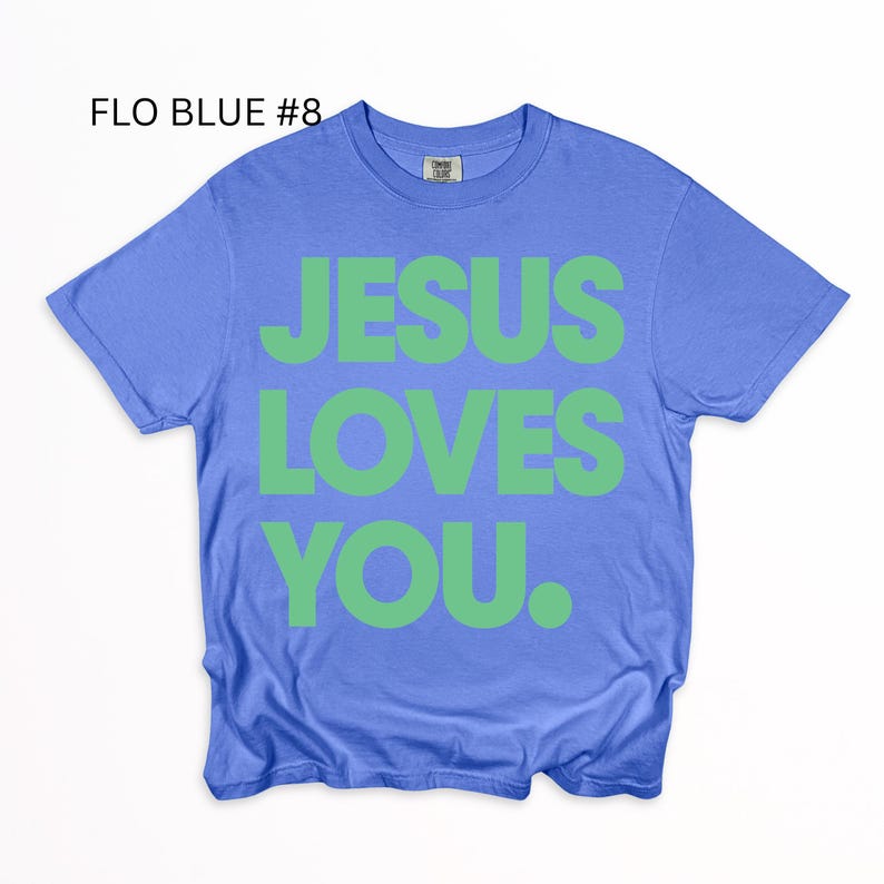 K&ouml;nnte beinhalten: Ein blaues T-Shirt mit dem Text "JESUS LOVES YOU" in gr&uuml;nen Blockbuchstaben. Das T-Shirt hat einen Rundhalsausschnitt. Der Text "FLO BLUE #8" befindet sich in der oberen linken Ecke.