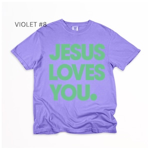 K&ouml;nnte beinhalten: Violettes T-Shirt mit dem Text "JESUS LOVES YOU" in gr&uuml;nen Blockbuchstaben. Das Shirt hat einen Rundhalsausschnitt und kurze &Auml;rmel. Der Text "VIOLET #8" befindet sich in der oberen linken Ecke.