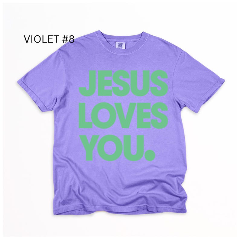 K&ouml;nnte beinhalten: Violettes T-Shirt mit dem Text "JESUS LOVES YOU" in gr&uuml;nen Blockbuchstaben. Das Shirt hat einen Rundhalsausschnitt und kurze &Auml;rmel. Der Text "VIOLET #8" befindet sich in der oberen linken Ecke.