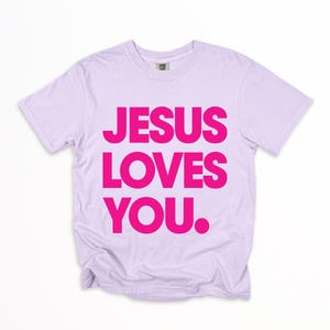K&ouml;nnte beinhalten: Ein helllilafarbenes T-Shirt mit dem Text "JESUS LOVES YOU" in leuchtend pinkfarbenen Blockbuchstaben. Das Shirt hat einen Rundhalsausschnitt und scheint aus einem weichen, bequemen Material zu sein. Der Text ist mittig auf der Vorderseite platziert.