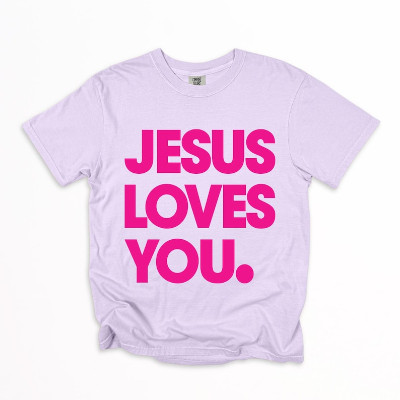 K&ouml;nnte beinhalten: Ein helllilafarbenes T-Shirt mit dem Text "JESUS LOVES YOU" in leuchtend pinkfarbenen Blockbuchstaben. Das Shirt hat einen Rundhalsausschnitt und scheint aus einem weichen, bequemen Material zu sein. Der Text ist mittig auf der Vorderseite platziert.