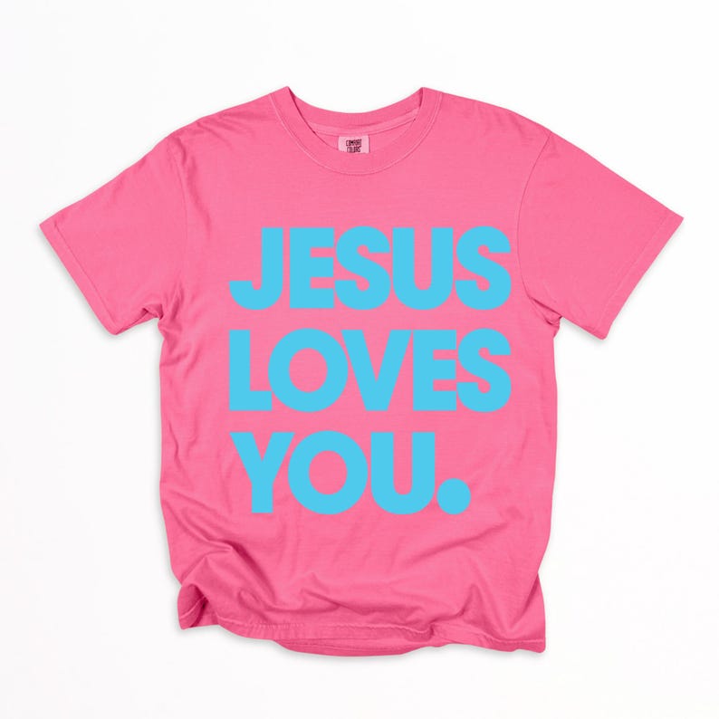 K&ouml;nnte beinhalten: Ein pinkfarbenes T-Shirt mit dem Text "JESUS LOVES YOU" in hellblauen Buchstaben. Das T-Shirt hat einen Rundhalsausschnitt und ist aus weichem Stoff gefertigt.