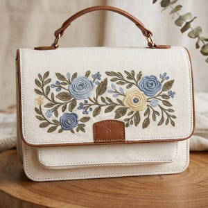 Puede incluir: Un bolso de mano de lona color crema con un patrón floral bordado de rosas azules y amarillas, hojas verdes y ribetes de piel sintética marrón. El bolso tiene un asa superior, una solapa frontal con una lengüeta marrón y una estructura rectangular.