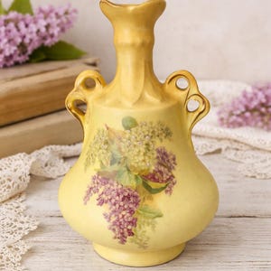 May include: A yellow ceramic vase with a floral design featuring purple and white flowers. The vase has two handles and a flared top. The background includes books and lace.