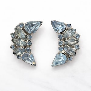 Puede incluir: Un par de pendientes de estilo vintage con una montura de metal plateado. Cada pendiente presenta una forma de media luna adornada con piedras de diferentes tamaños, azul claro y transparentes. Las piedras están dispuestas para crear un diseño brillante y elegante.
