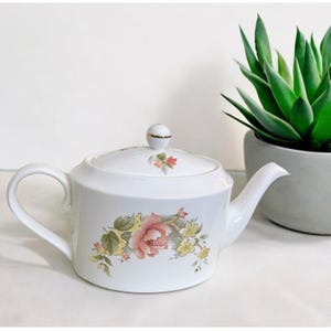 May include: White ceramic teapot with a floral design in pink, yellow, and green. The teapot has a curved handle and spout, and a lid with a gold-trimmed knob. The floral pattern is on the side of the teapot. A decorative item for the kitchen.