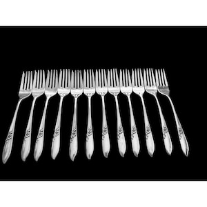 Op de afbeelding: Een verzameling zilveren vorken tegen een zwarte achtergrond. Elke vork heeft een bloemmotief op de steel. De tanden zijn gelijkmatig verdeeld en de vorken zijn lichtjes schuin geplaatst, wat een aantrekkelijke presentatie creëert. Ideaal voor een formeel diner.