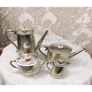 May include: A four-piece silver tea set comprising a teapot, creamer, sugar bowl, and small pitcher. Each item features a floral design and a light-colored knob on the lid. The set is presented on a white tablecloth, ideal for serving tea.
