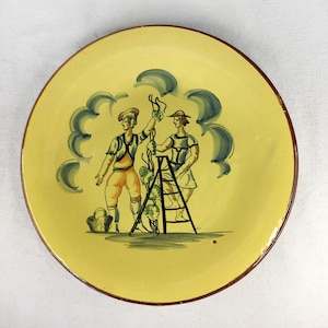 May include: A yellow ceramic plate with a brown rim features a hand-painted scene of two people harvesting grapes. The scene includes a ladder, a basket, and clouds in the background.