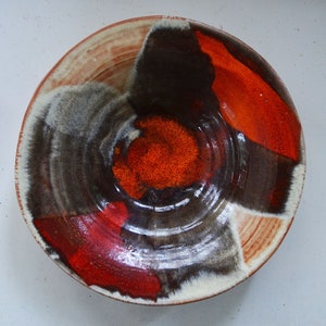 Puede incluir: Un cuenco de cerámica con un esmalte marrón oscuro y acentos rojos y blancos. El cuenco tiene una forma circular y un borde ligeramente elevado.