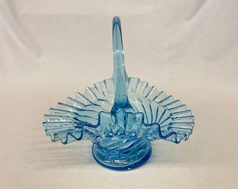 Puede incluir: Una cesta de cristal azul claro con asa. La cesta tiene un borde con volantes y una base en espiral. El asa es arqueada y se conecta al cuerpo de la cesta. El cristal es translúcido.