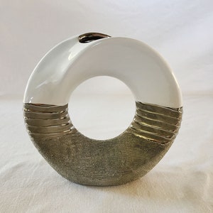 May include: A white ceramic vase with a silver and gold textured design. The vase is shaped like a ring and has a wide opening at the top.