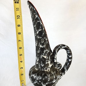 May include: A black and white ceramic vase with a long, thin neck and a handle. The vase has a speckled glaze and a red rim.