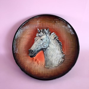 Puede incluir: Un plato de pared redondo de cerámica con una escultura en relieve de cabeza de caballo blanca en el centro. El plato tiene un fondo marrón y naranja con un patrón de cuadrícula.