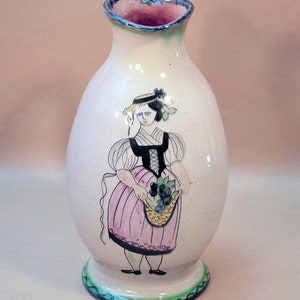Op de afbeelding: Een witte keramische kan met een roze en groene handvat. De kan is versierd met een handgeschilderd beeld van een vrouw in een roze jurk en een strohoed die een mand met druiven vasthoudt.