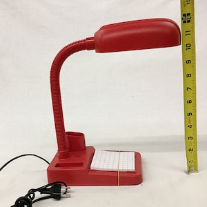 Peut inclure: Une lampe de bureau rouge avec un col flexible et un porte-crayons intégré. La lampe a un bloc-notes quadrillé blanc fixé à la base.