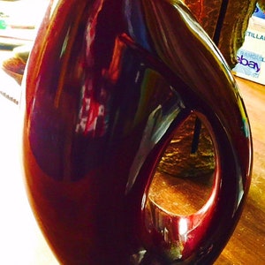 May include: A dark red ceramic vase with a unique, abstract shape and a cutout on the side. The vase has a glossy finish and reflects light.