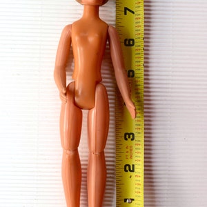 May include: A nude plastic doll with articulated limbs, standing next to a yellow measuring tape. The doll is approximately 10 inches tall.