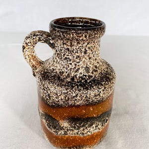 May include: A small, vintage ceramic pitcher with a handle. The pitcher features a textured, speckled surface in shades of brown, black, and cream. Horizontal bands of brown and black encircle the base. The opening is dark brown.