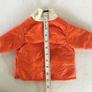May include: An orange quilted jacket with a cream-colored ribbed collar. A measuring tape is placed next to the jacket. The jacket appears to be approximately 12 inches in length. The jacket has long sleeves and a quilted design.