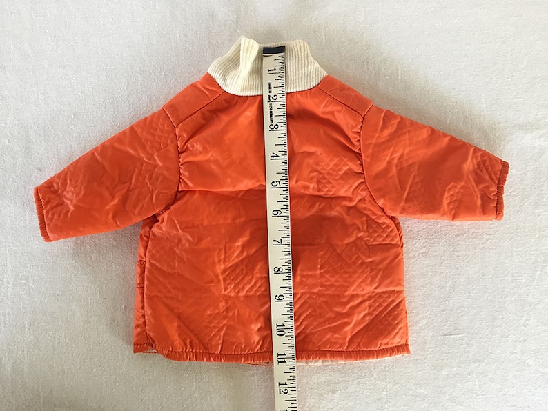 May include: An orange quilted jacket with a cream-colored ribbed collar. A measuring tape is placed next to the jacket. The jacket appears to be approximately 12 inches in length. The jacket has long sleeves and a quilted design.