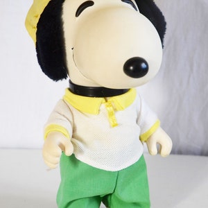 Pode incluir: Um brinquedo de pelúcia do Snoopy vestindo um boné de beisebol amarelo, uma camisa branca com gola amarela e calças verdes.