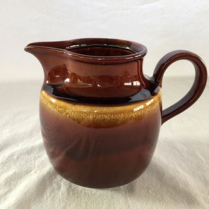 Puede incluir: Una jarra de crema de cerámica marrón con un acabado esmaltado. La jarra tiene un cuerpo redondeado y un asa.